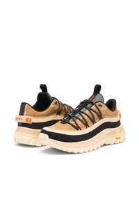 Beiget ja mustat urheilukengät, joissa paksu valkoinen pohja, nauhat, joustavat lenkit, teksturoidut materiaalit ja näkyvä logo kannassa.