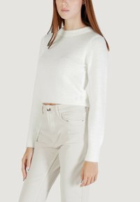 Donna che indossa un maglione corto a maniche lunghe bianco e morbido, insieme a pantaloni beige chiaro a vita alta, contro uno sfondo semplice.