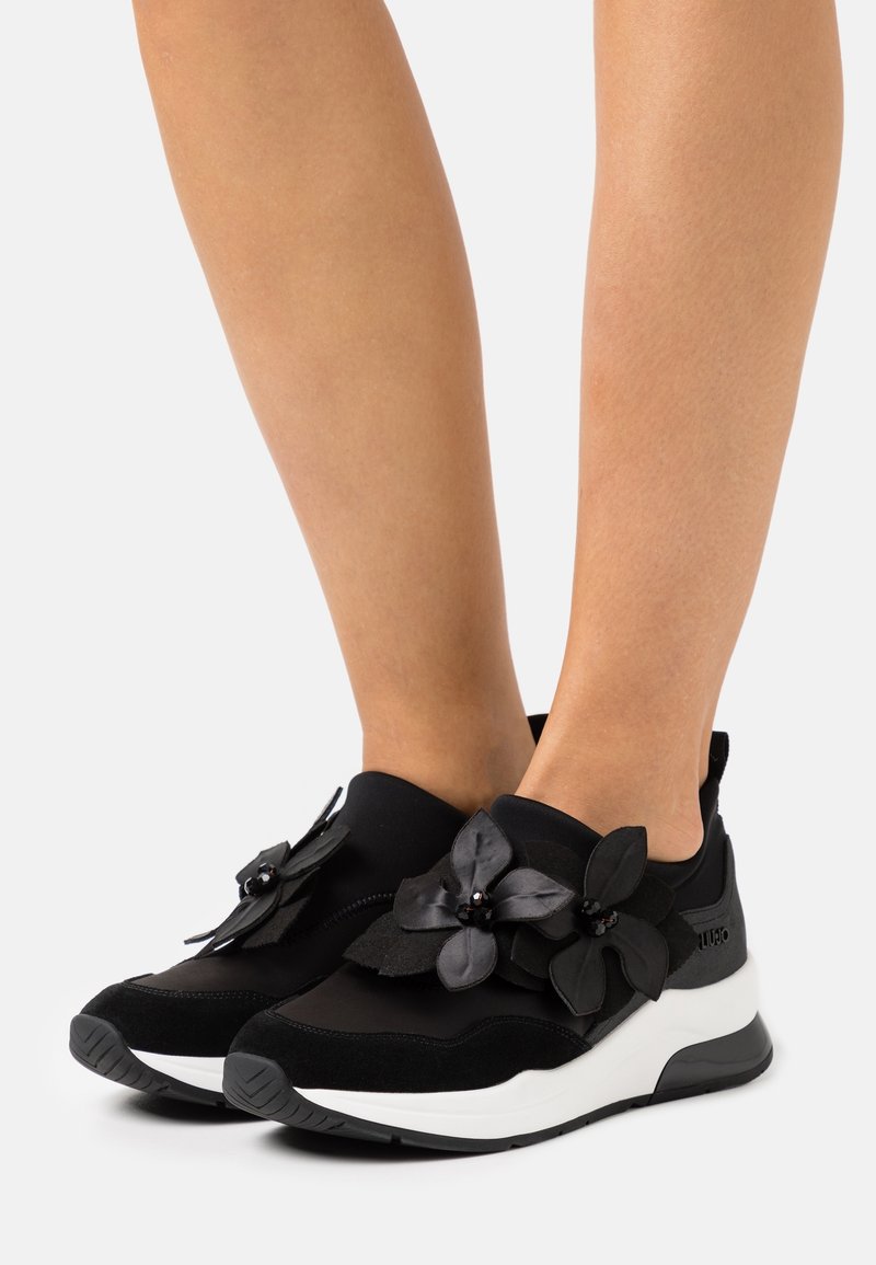 Schwarze Sneaker mit einem Obermaterial aus Wildleder, verziert mit großen schwarzen Blumenapplikationen und einer weißen Gummisohle. Texturiertes Design mit moderner Silhouette.