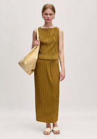 Khaki plooijurk met een ronde halslijn, trekkoord in de taille en bijpassende beige tas. Model draagt een paar crèmekleurige sandaaltjes en staat tegen een neutrale achtergrond.
