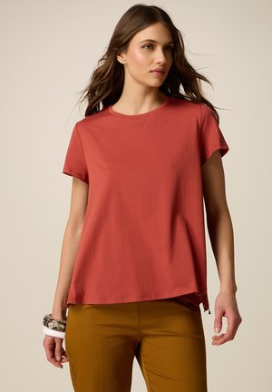 Donna con i capelli lunghi che indossa una maglietta a maniche corte color ruggine e pantaloni color senape, accessoriata con diversi braccialetti al polso sinistro.