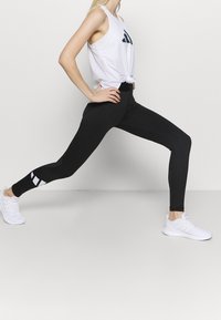 Femme en baskets blanches, leggings noirs et débardeur blanc s'étirant dans une pose de fente avant devant un fond uni.