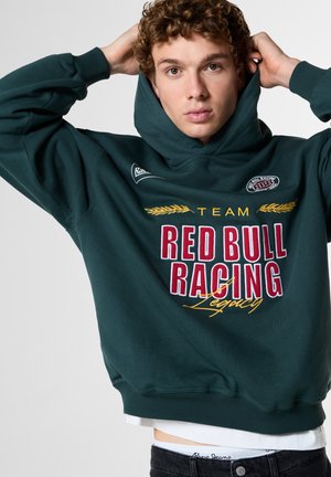 Jonge man met krullend haar die een donkergroene Red Bull Racing hoodie draagt en de capuchon over zijn hoofd trekt tegen een effen achtergrond.