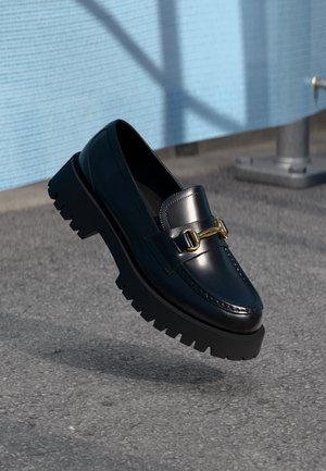 Navy leather loafers with a chunky black outsole, featuring a decorative gold hardware accent and stitching detail on the upper.