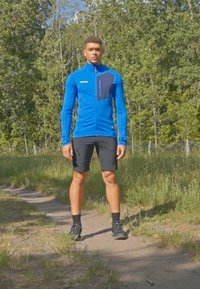 Blue zip-up jacket with black accents, long sleeves, and textured fabric; paired with black shorts and athletic shoes on a grassy path.