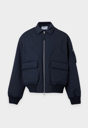 Jeune homme portant une casquette jaune pâle, une veste bomber noire, une chemise rose, un jean bleu clair et des baskets beiges, debout avec une main dans la poche.