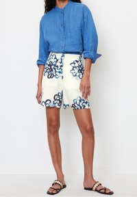 Chemise en lin bleue à boutons associée à un short crème orné de motifs floraux bleus. Sandales plates noires avec détails festonnés.