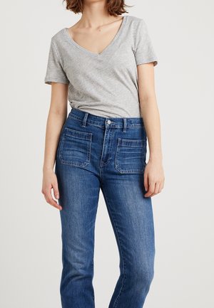 Kvinde iført en grå V-hals kortærmet skjorte stukket ned i højtaljede blå jeans med frontlomme, stående mod en hvid baggrund.