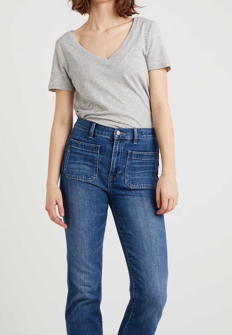 Mujer con camiseta de cuello en V de manga corta en gris jaspeado, metida dentro de jeans azul de talle alto con bolsillos frontales, de pie frente a un fondo blanco.