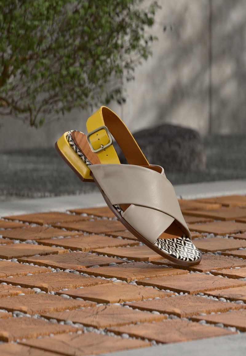 Μπεζ δερμάτινο σανδάλι με σταυρωτά λουράκια, κίτρινη αγκράφα στον αστράγαλο και ασπρόμαυρη επένδυση με σχέδιο, τοποθετημένο σε ξύλινη επιφάνεια με υφή.