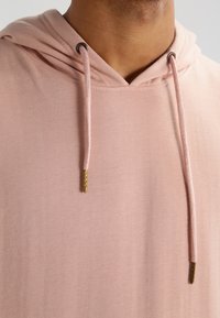 Felpa in cotone rosa chiaro con cappuccio regolabile, caratterizzata da punte metalliche dorate sui lacci. Texture morbida, design casual.