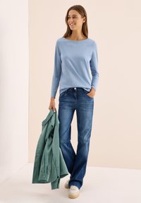 Femme souriante, portant une chemise à manches longues bleu clair et un jean, tenant une veste en velours côtelé verte à une main, se tenant devant un fond neutre.