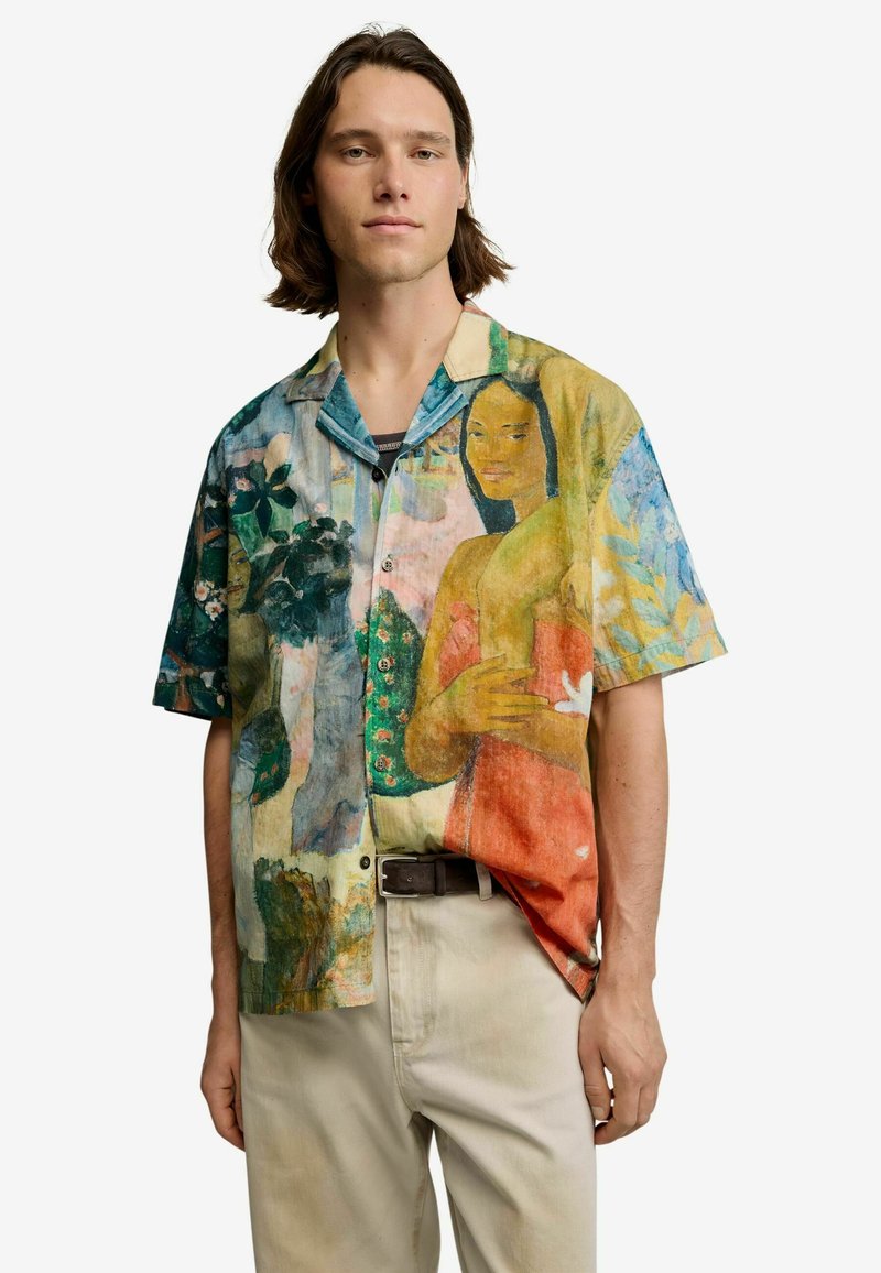 Young man wearing a short-sleeve button-up shirt with a colorful print of a woman in a red dress and abstract nature elements, paired with light pants.