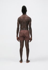 Brązowe krótkie spodenki kąpielowe z lekkiego materiału, z elastycznym paskiem i gładkim przodem, widziane od tyłu, na neutralnym tle.