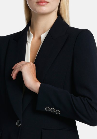 Blazer marine à la coupe ajustée, avec des revers pointus, des poignets à quatre boutons, et un tissu lisse. Une blouse de couleur claire est visible en dessous.