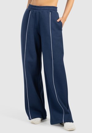 Persoon draagt donkerblauwe wijde broek met witte verticale strepen en witte schoenen, staat met één hand in de zak.