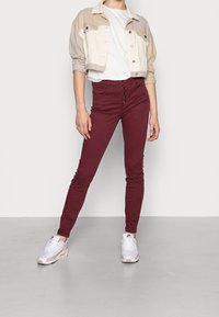 Jean skinny bordeaux avec bouton sur le devant ; associé à une veste cropped bicolore beige et crème ; t-shirt blanc en dessous ; stylé avec des baskets.