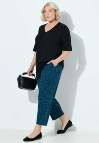 Schwarzes V-Ausschnitt-T-Shirt, tealfarbene Leopardenmuster-Hose, schwarze Ballettschuhe, hält eine kleine schwarze Handtasche mit einem goldenen Griff.