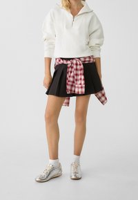 Sweat-shirt blanc à col zippé, jupe plissée noire, chemise flanelle rouge à carreaux nouée à la taille, baskets argentées et chaussettes blanches montant à la cheville.