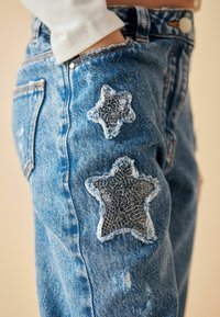 Jeans de mezclilla con un lavado claro que presentan dos parches en forma de estrella en negro y plata, cosidos con bordes deshilachados para añadir textura.