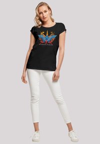 Svart t-skjorte med kort erm som har en fargerik "Wonder Woman" logo med blå og røde detaljer, kombinert med hvite skinny jeans og hvite sneakers.