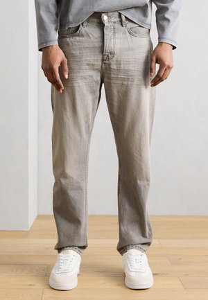 Personne portant un jean gris à jambes droites et des baskets blanches, debout sur un sol en bois clair, sur un fond clair uni.