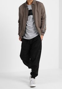 Muž v taupe bomber bundě s bílými pruhy, šedém tričku s potiskem, černých cargo kalhotách a bílých teniskách, stojící se zkříženýma rukama v kapsách.