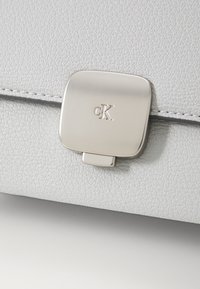 Hvit teksturert skinnveske med et sølvfarget firkantet lås med cK-logoen, som fremhever minimalistisk design og kvalitetsbeslag.