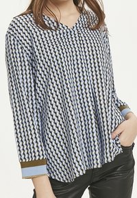 Kvinna klädd i en långärmad blus med geometriskt mönster och V-ringning samt svarta byxor, med handen vilande i fickan.