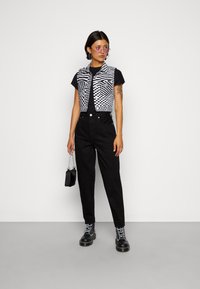 Nuori henkilö, jolla on päällä mustat housut, musta T-paita, mustavalkoinen ruudullinen liivi, mustat saappaat, vaaleanpunaiset aurinkolasit ja hän pitää pientä mustaa laukkua.