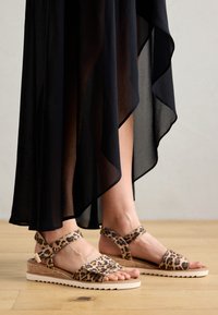 Leopard print sandals with a cork base, adjustable ankle strap, and a light-colored rubber sole. Paired with a flowing black dress.