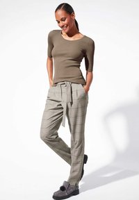 Olijfgroene geribbelde top met korte mouwen gecombineerd met grijs geruite high-waisted broek met een strikceintuur. Model draagt grijze schoenen met decoratieve accenten.