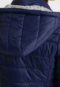 Chaqueta acolchada azul marino con textura acolchada, que cuenta con un forro de capucha gris adornado con pequeñas estrellas. Diseñada con un ajuste relajado.