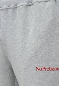 Κοντινή εικόνα γκρι υφάσματος φόρμας με κόκκινη κεντημένη επιγραφή "No Problemo" κοντά στην περιοχή της τσέπης.