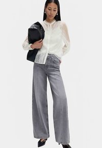 Hvid blondebluse med lange ærmer, kombineret med højtaljede, grå viddebukser i denim. Modellen holder en sort shoppertaske og har sorte, slanke hæle på.