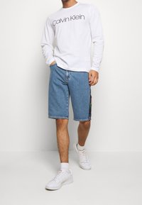 White long-sleeve shirt with "Calvin Klein" logo, paired with light blue denim shorts featuring multicolored side accents; white shoes.