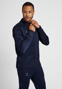 Veste de sport navy avec un col haut, fermeture éclair et logo sur la poitrine. Fabriquée en tissu lisse et avec une coupe structurée.
