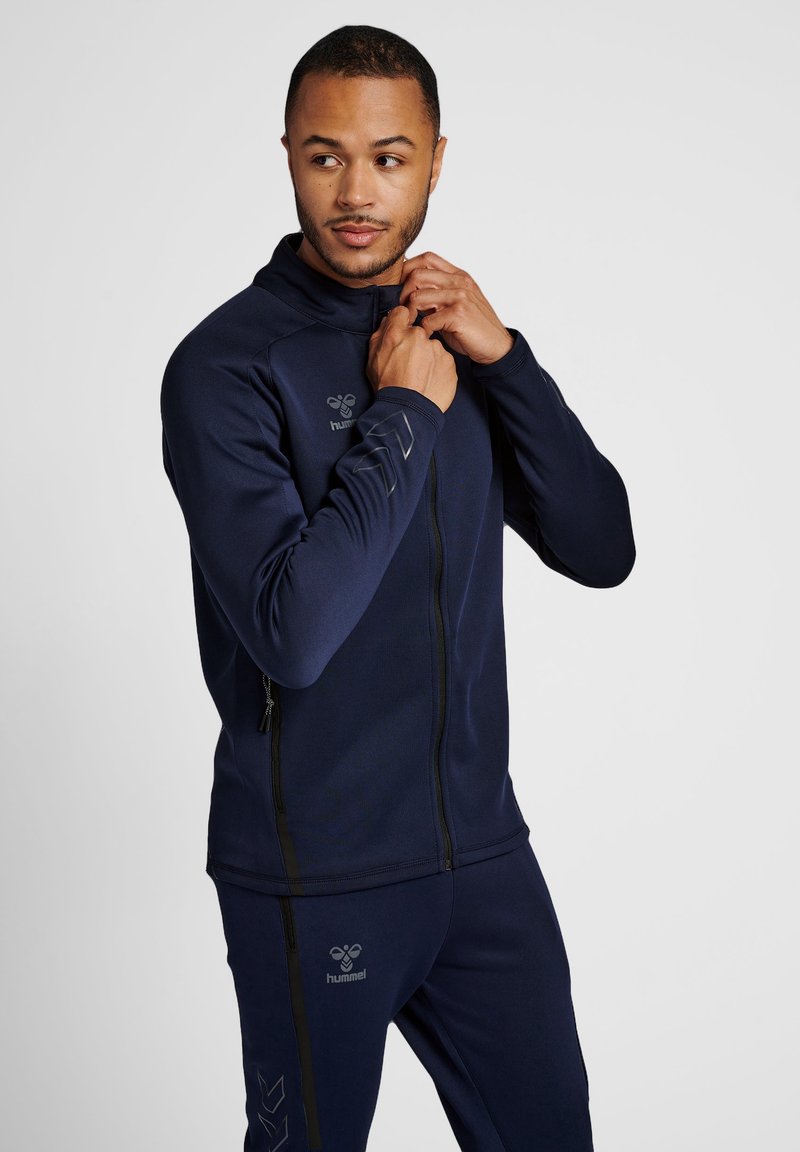 Veste de sport navy avec un col haut, fermeture éclair et logo sur la poitrine. Fabriquée en tissu lisse et avec une coupe structurée.