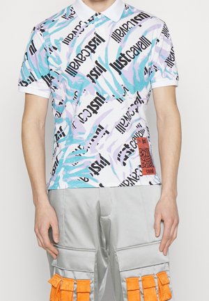 Mann trägt ein weißes Poloshirt mit dem Schriftzug "Just Cavalli" in Schwarz und Pastellblau, kombiniert mit hellgrauen Hosen mit orangen Trägern.