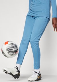 Gaiši zili sporta bikses ar ribotu tekstūru, kopā ar melniem un baltiem amerikāņu futbola zābakiem. Netālu ir kustībā rakstains futbola bumbas.