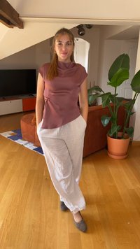Priliehavý mauve top s vysokým golierom a detailom na boku, skombinovaný s voľnými bielymi ľanovými nohavicami a šedými plochými topánkami, umiestnený v obývacej miestnosti.