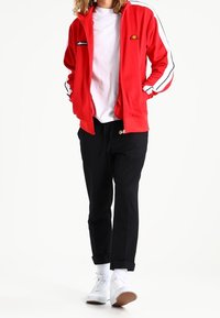 Rød lynlåstrøje med hvide sidelister og logo, kombineret med sorte tilspidsede bukser og hvide sneakers. Hvid t-shirt under.