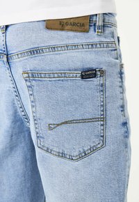 Vaqueros de denim de color azul claro con un diseño de cinco bolsillos, con costuras amarillas, un parche de marca de cuero en la cintura y una etiqueta azul marino en el bolsillo trasero.