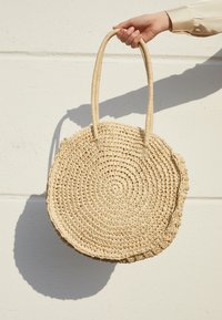 Rund, vävd stråväska med två texturerade, beigefärgade handtag. Har ett spiralmönster och en något rufsig kant för extra detalj.