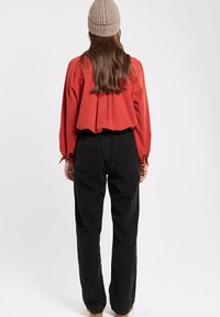 Blouse rouge cropped avec épaules tombantes et plis au dos, associée à un pantalon noir à jambes larges avec deux poches arrière. Bonnet en tricot beige.
