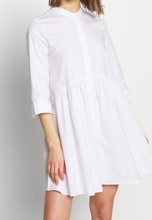 Žena v bílé šatové košilové sukni se stahováním v pase a tříčtvrtečními rukávy, stojící na jednoduchém světlém pozadí.