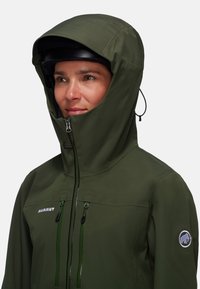 Giacca impermeabile verde con colletto alto e grande cappuccio, dotata di due tasche frontali con zip, coulisse elastica e patch con logo sulla manica.