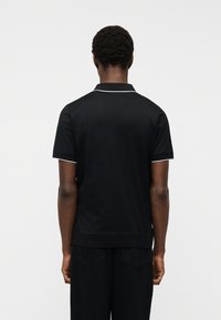 Άνδρας με κοντά σγουρά μαλλιά στέκεται με την πλάτη στραμμένη, φορώντας μαύρο πουκάμισο με κοντά μανίκια και λευκή επένδυση στον γιακά και στις άκρες των μανικιών.