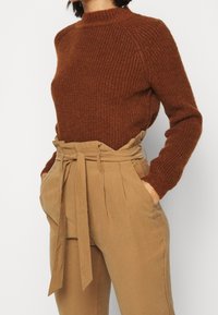 Brun texturerad stickad tröja med hög halsringning, parat med beige högmidjade byxor med knytbälte och veckdetaljer.