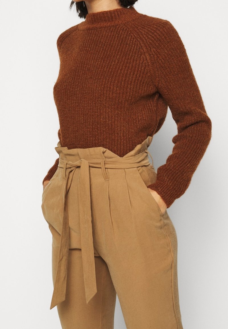 Brun texturerad stickad tröja med hög halsringning, parat med beige högmidjade byxor med knytbälte och veckdetaljer.